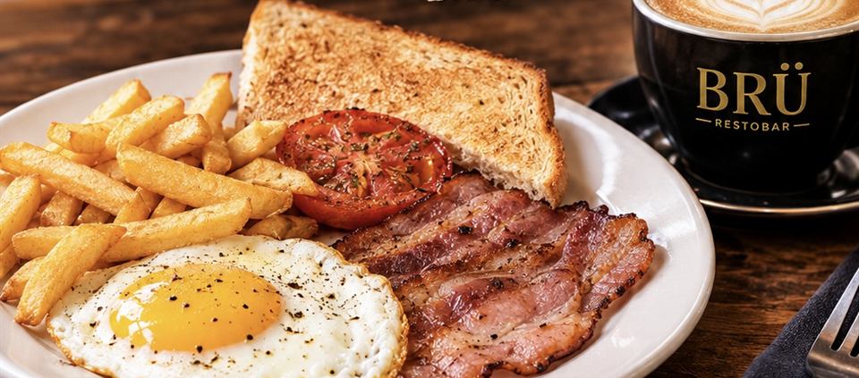 Breakfast plate with a fried egg, bacon, chips, grilled tomato and toast, with a BRÜ cappuccino